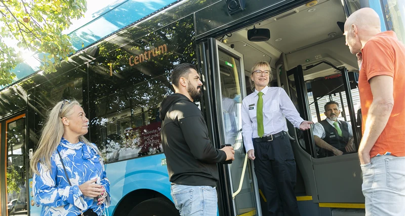 Bussförare står i öppningen till en buss och hälsar passagerare välkomna ombord.