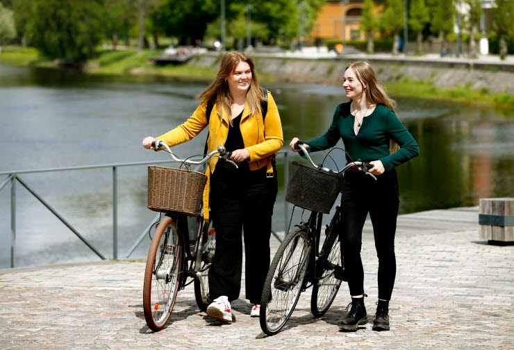 Två tjejer går med sina cyklar med Klarälven i bakgrunden.