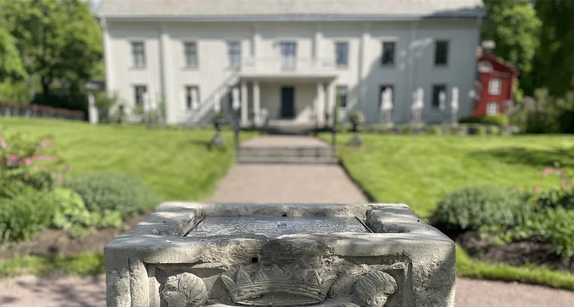 Stenbrunn i förgrunden med park och vit byggnad i bakgrunden