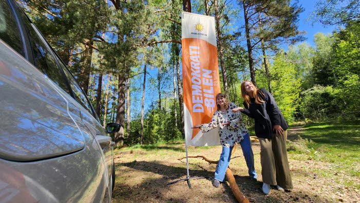 Miriam och Malin står i sommarlandskap bredvid en bil och framför en strandflagga med texten "Demokratibilen på turné".
