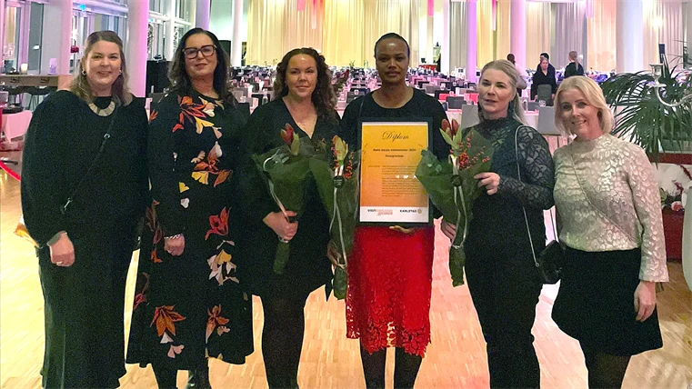 Sex kvinnor står en på en rad framför fotografen. En av kvinnorna i mitten håller i ett diplom.