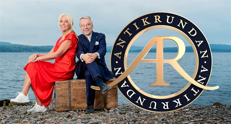en kvinna i röd klänning och en man i kostym sitter på en trälåda på en strand med vatten i bakgrunden