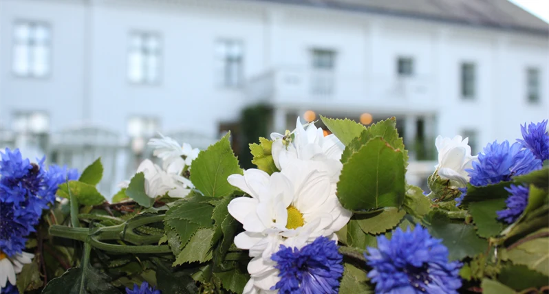 Herrgård i fond med del av blomsterprydd midsommarstång i förgrund.