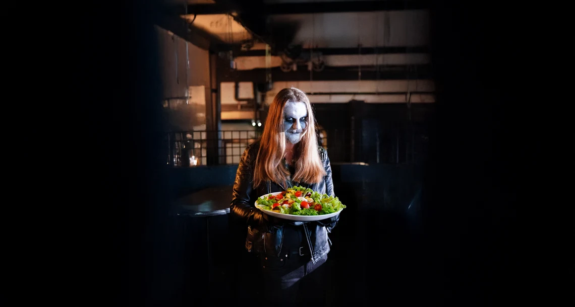 En person med långt rött hår och ansiktsmålning i svart och vitt, står i ett mörkt industriellt rum och håller en tallrik med färgglad sallad. Personen bär svarta kläder och ser ner på maten med allvarlig min.