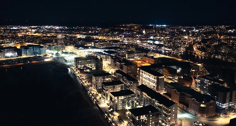 En flygbild tagen nattetid över en stad med ljus från fönster och gatubelysning.