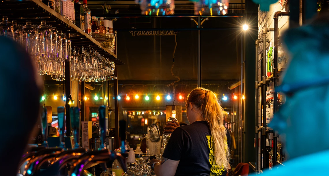 Två personer sitter i förgrunden av en bar där en bartender står och häller upp något.