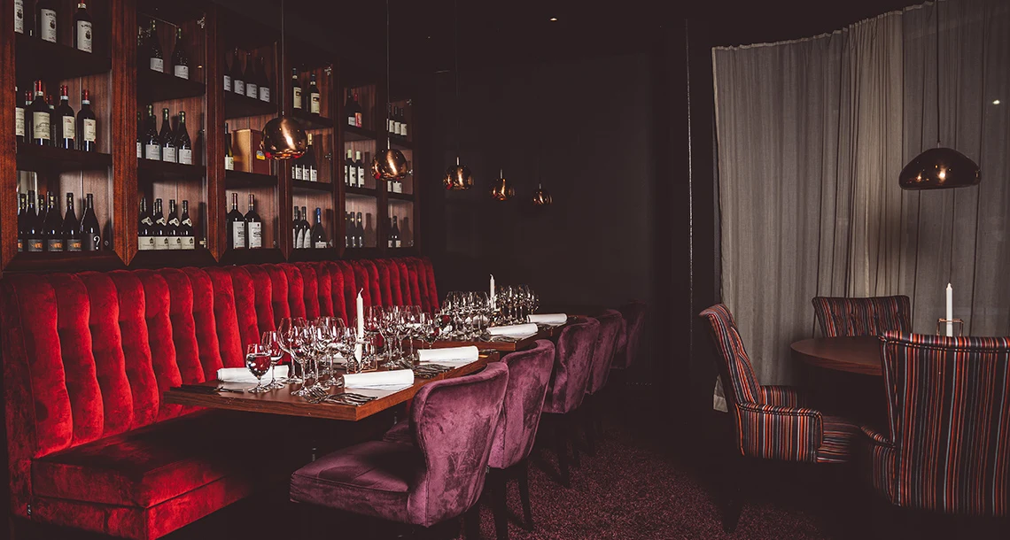 En stilig soffa i röd plysch med dukade bord framför och hyllor med vinflaskor bakom.