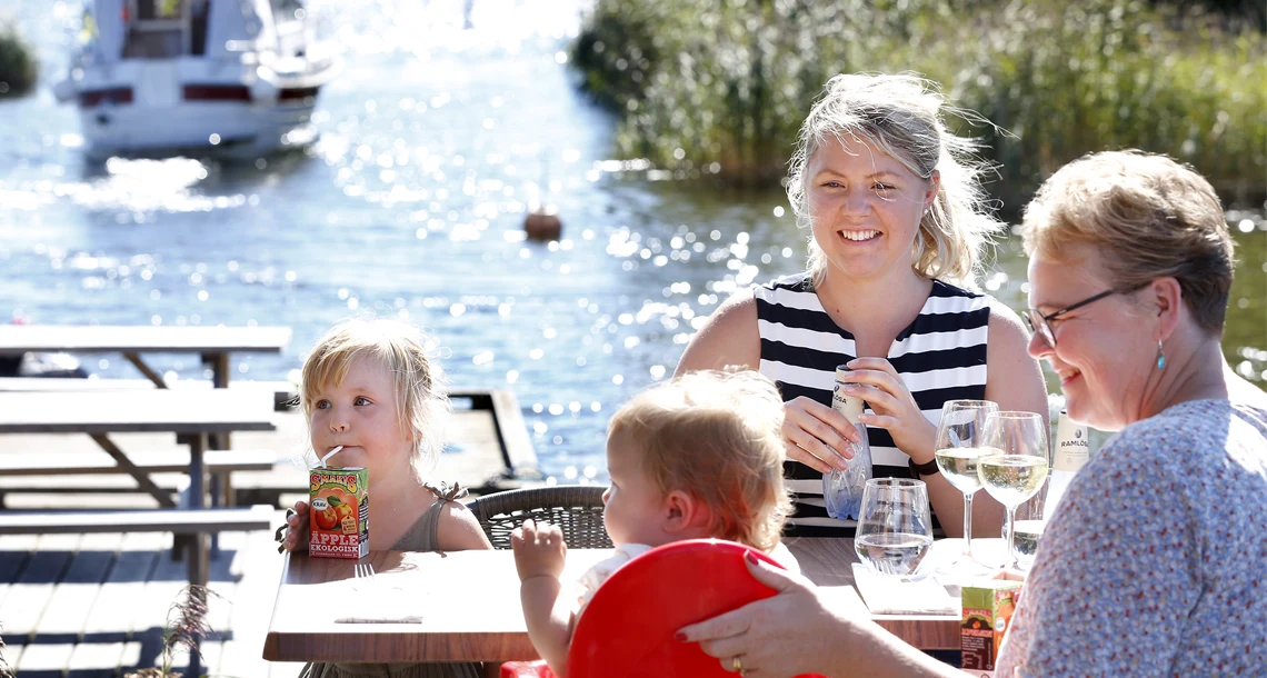 En kvinna, en äldre dam och två barn njuter av dryck vid ett bord. I bakgrunden ses en båt i vattnet