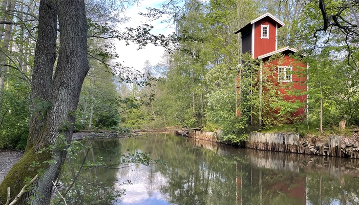På bilden syns det röda elevatorhuset med vita knutar som speglar i en vattenyta. Huset är omgivet av träd och grönska, och himlen syns också i reflektionen