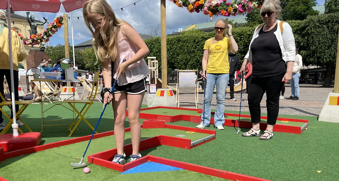 Mormor, tonåring och barn spelar minigolf. 