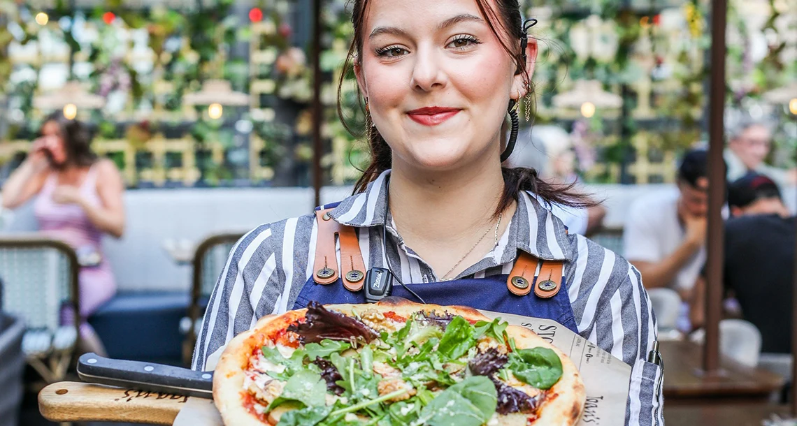 En tjej i servitörskläder håller upp en pizza på en träbricka