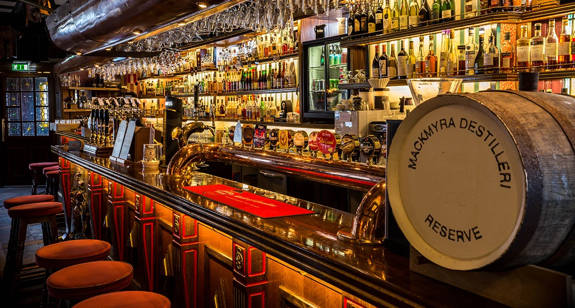 En bardisk i mörka färger framför flera rader av flaskor uppställda i hyllor