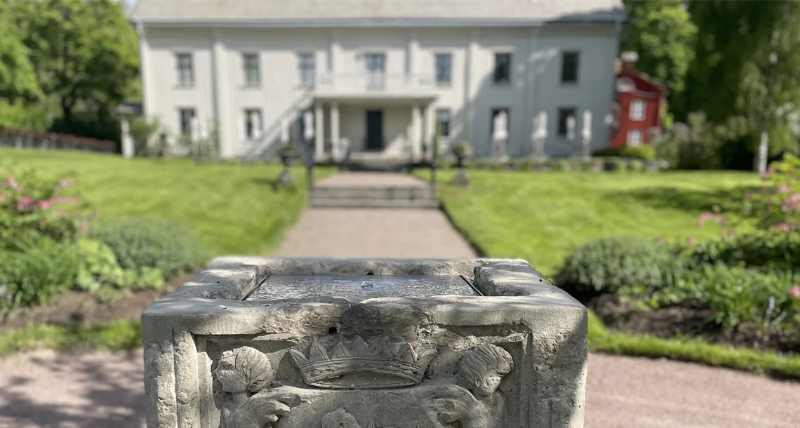 Stenbrunn i förgrunden med park och vit byggnad i bakgrunden