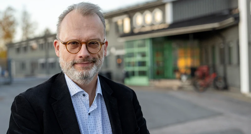 Porträttbild av en person i mörk kavaj och ljus skjorta utomhus. I bakgrunden syns en byggnad med stora fönster och grön entré.