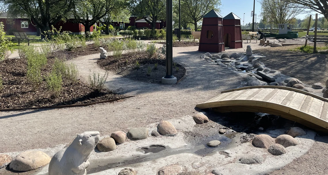 Grusgångar i en park men liten träbro över ett vattendrag och en stenbäver. Lekhus i form av dykdalber i bakgrunden