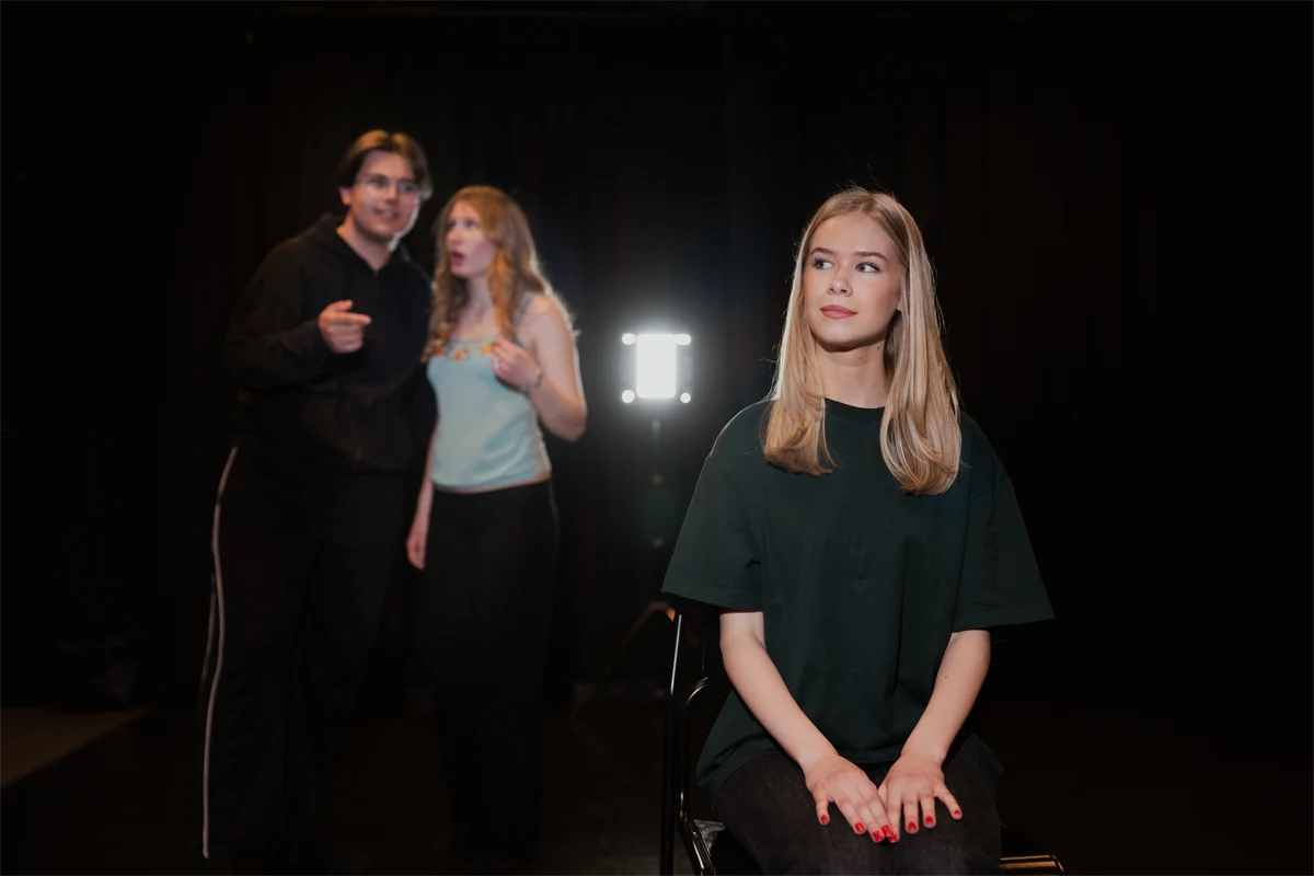 Gymnasieelever spelar teater i ett mörkt rum med strålkastare.