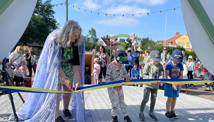 En kvinna med glittrig cape klipper en Sverigeband tillsammans med tre barn i keps.