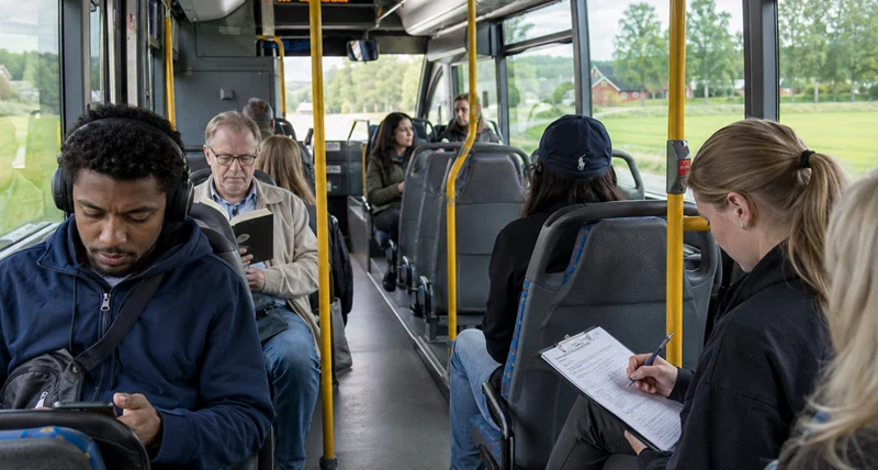 insida av buss i färd. Flera passagerare sitter. Utanför grönskande landsbygd