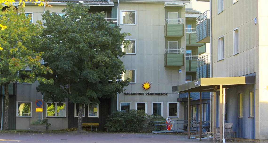 Vy över entrén till Hagaborgs trygghetsboende. På stenhuset pryder karlstadsolen med texten "Hagaborgs vårdboende". Frramför byggnanden står träd som skiftar i olika gröna nyanser.