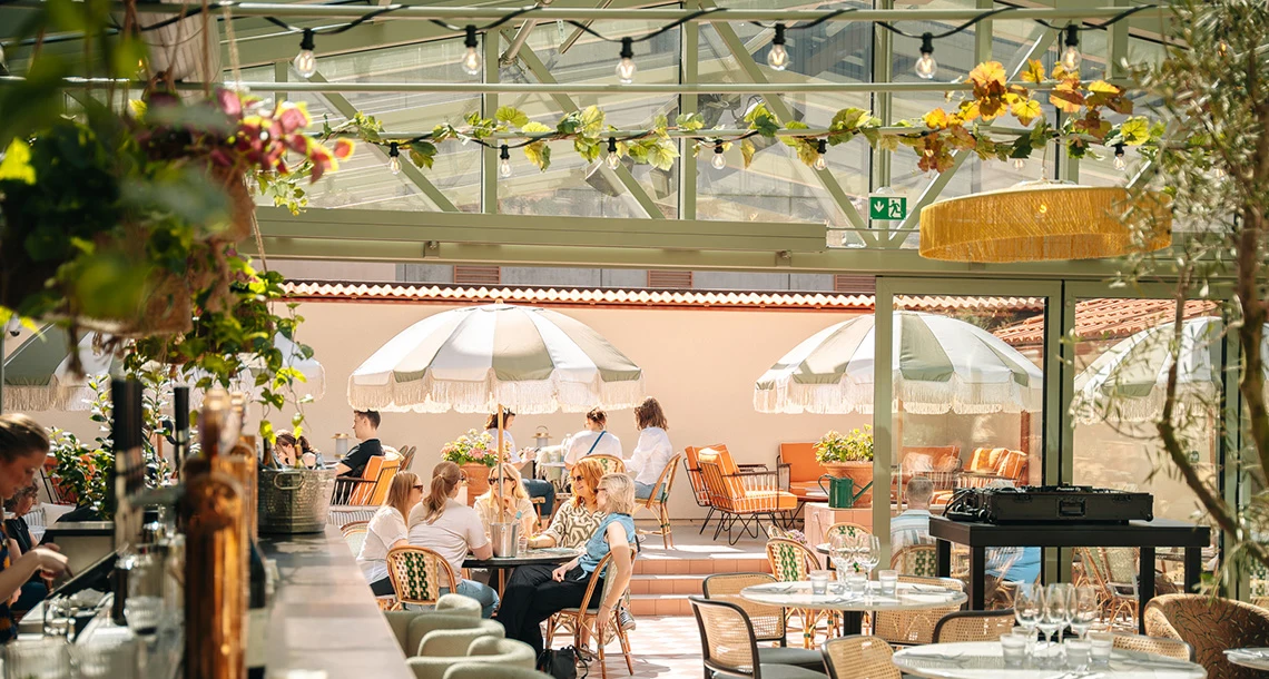 En bardisk i förgrunden bredvid flera bord och i bakgrunden sitter människor och äter och dricker