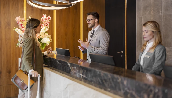 Två receptionister jobbar i en reception. Den ena hjälper en gäst att checka in.