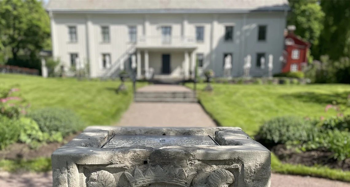 Stenbrunn i förgrunden med park och vit byggnad i bakgrunden
