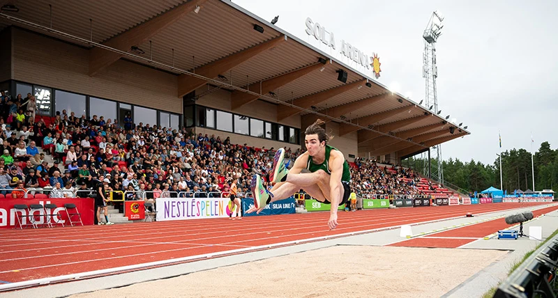 En man i sportkläder hoppar längdhopp och  bakgrunden finns en stor läktare med publik.