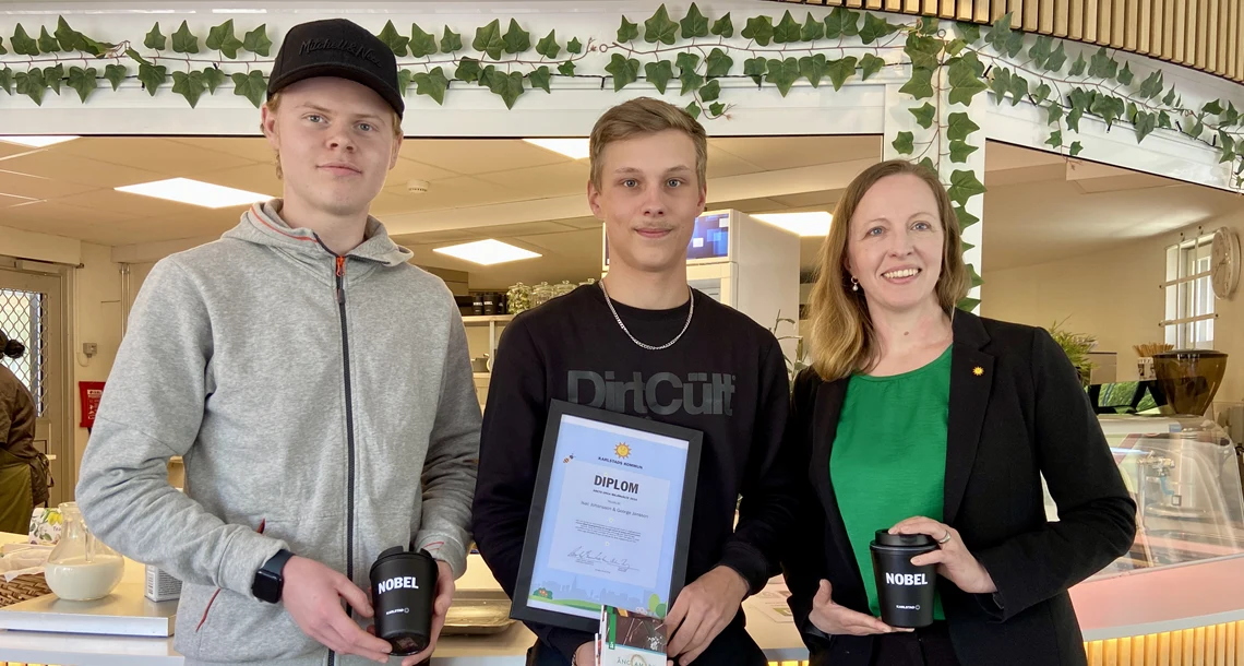 Bild på när Isac och Georg, som blev årets unga miljöhjältar 2025 får sitt diplom av miljönämndens ordförande Cecilia. De tre står på rad och håller upp varsin svart kaffemugg med texten "Nobel" på som de har fått pris för. Samt ett inramat diplom.
