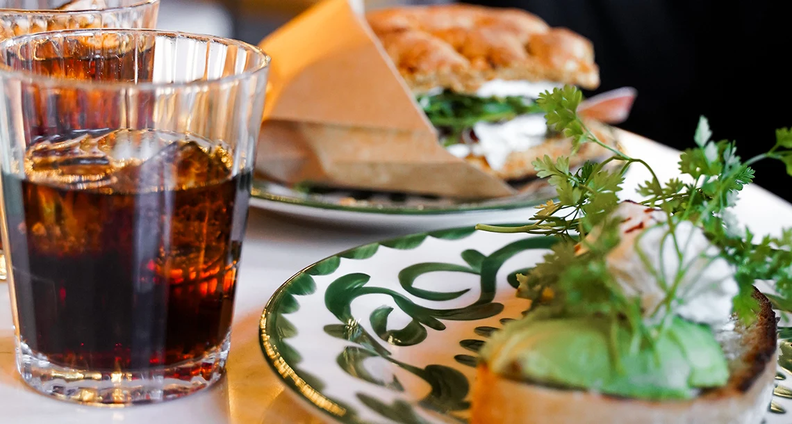 En glas med brun dryck och ett fat med en toast med grönt på.