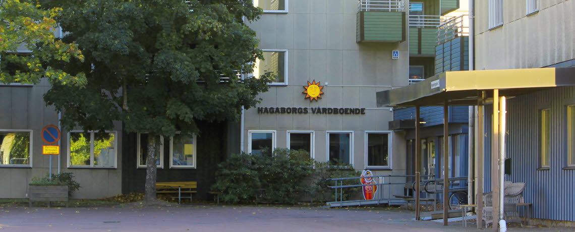 Vy över entrén till Hagaborgs trygghetsboende. På stenhuset pryder karlstadsolen med texten "Hagaborgs vårdboende". Frramför byggnanden står träd som skiftar i olika gröna nyanser.