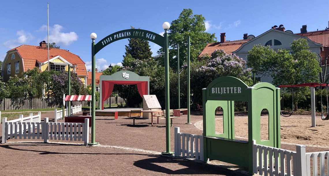 Bakom en vitt staket syns en liten scen med trädraperier och en biljettlucka.