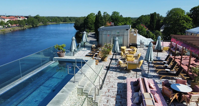 En liten utomhuspool och flera solstolar och parasoller på ett högt hustak med vatten och träd i bakgrunden.