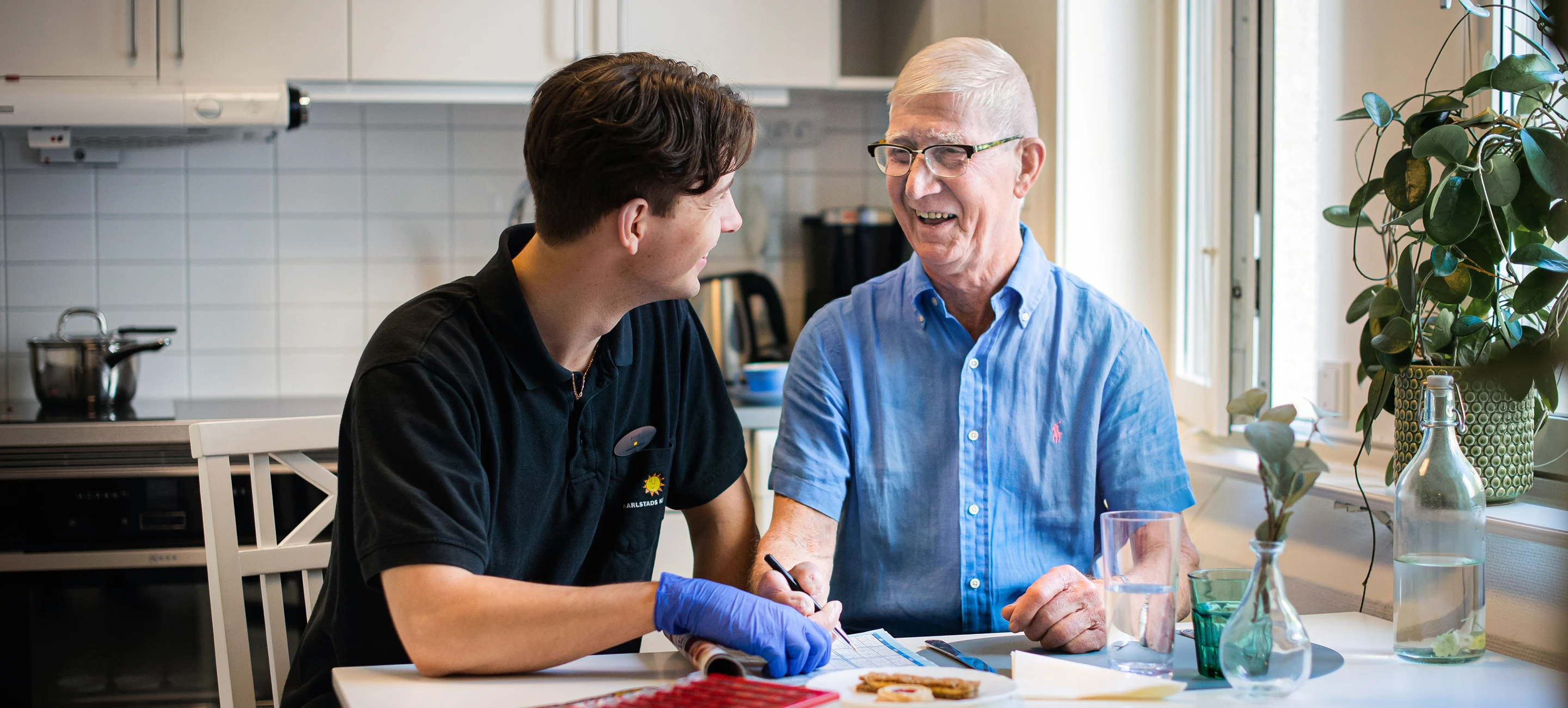 En äldre man som sitter vid ett köksbord med en vårdpersonal