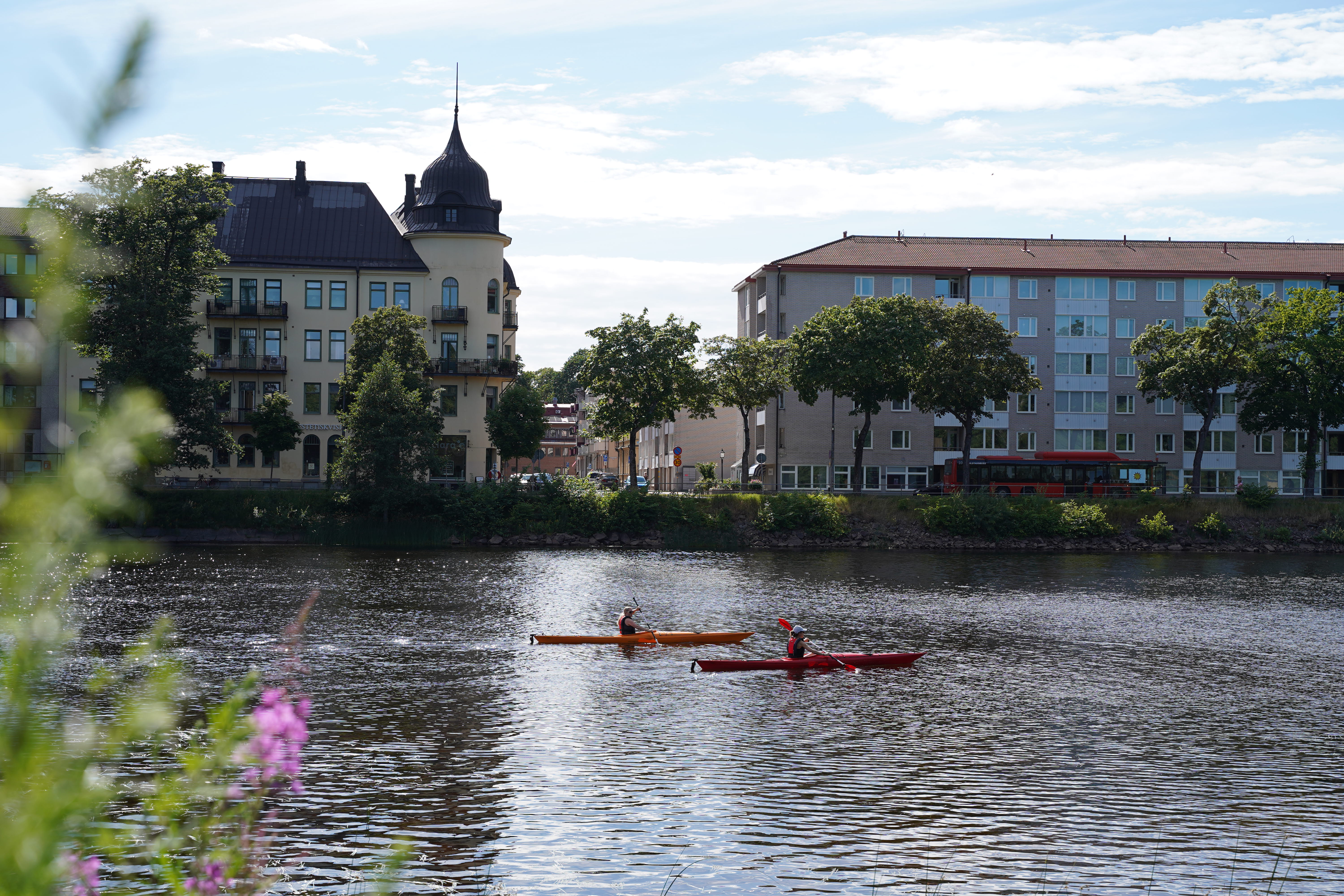 En vybild över Klarälven där ett par människor paddlar kanot. Bortom älven syns byggnader i Karlstad