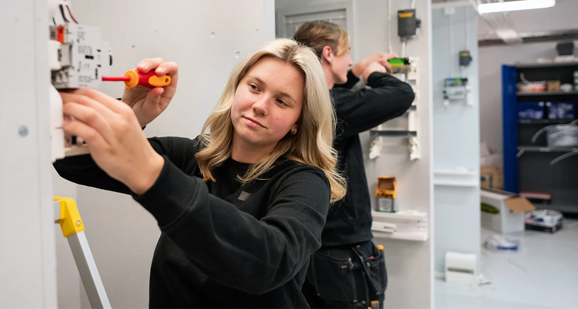 En tjej och en kille står vid var sin vägg i verkstadsmiljö och installerar el.