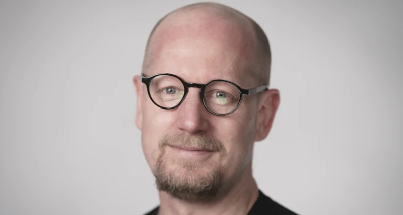 Porträttbild av en person med rakad skalle och runda glasögon som är vänd mot kameran. Personen har kort skägg och bär en mörk tröja. Bakgrunden är ljus och jämn utan tydliga detaljer, och bilden är mjukt belyst med fokus på ansiktet.