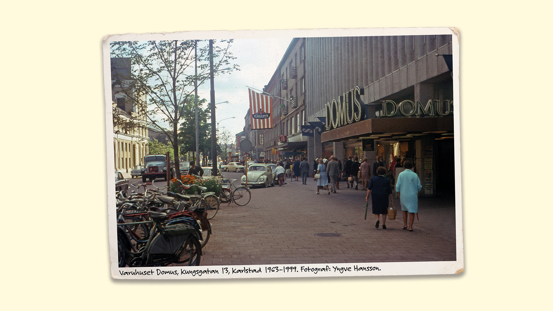 Gammalt foto på Kungsgatan 13 i Karlstad. I bilden ser man människor och gamla bilar på gatorna. Man ser också en stor skylt där de står Domus.