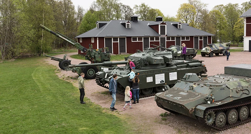 Flera olika militärfordon står uppställda på en grusplan med barnfamiljer och barn som klättrar på fordonen.