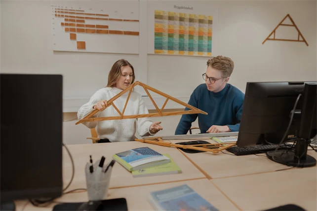 Två personer i ett klassrum tittar på en konstruktion i trä.