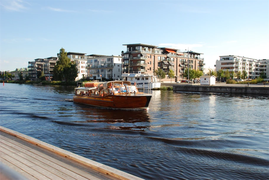 En mindre passagerarbåt i trä åker förbi en brygga och i bakgrunden finns ett bostadsområde med våningshus.