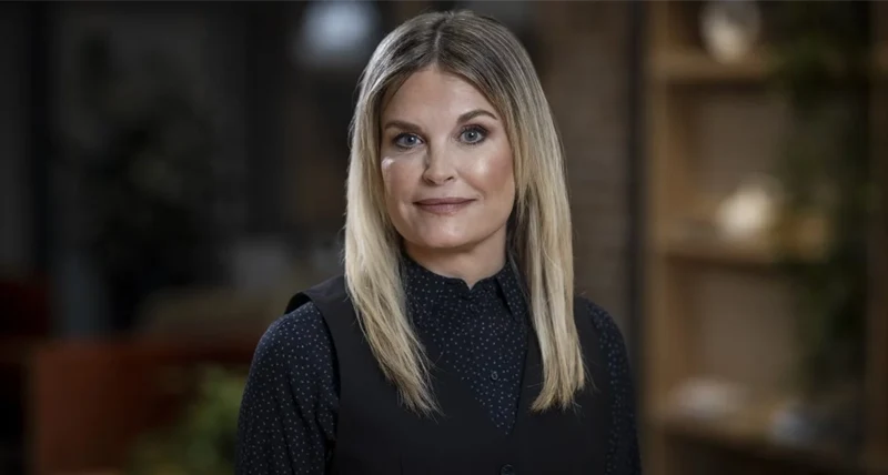 Porträttbild av en person med axellångt ljust hår som står vänd mot kameran. Personen bär en mörk långärmad skjorta med små prickar och en svart väst ovanpå. Bakgrunden är inomhus och suddig, med hyllor och dekorativa föremål i varma färgtoner.