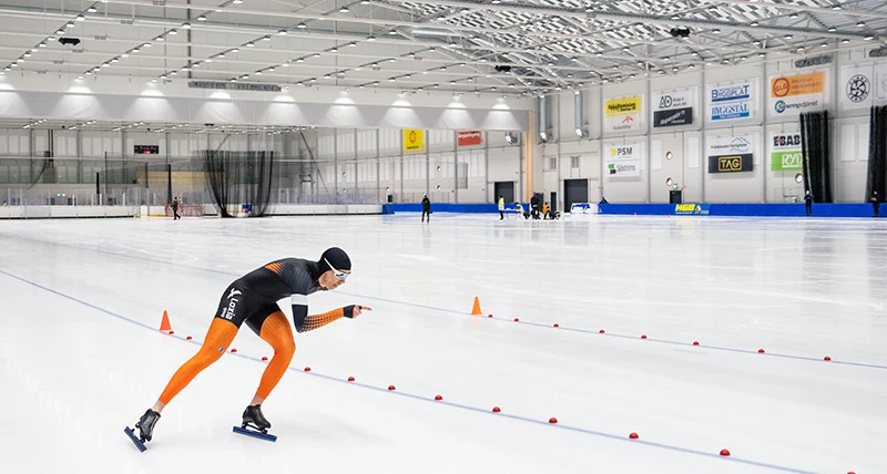 En man i svarta och orangea lycrakläder åker skridskor i en stor inomhushall