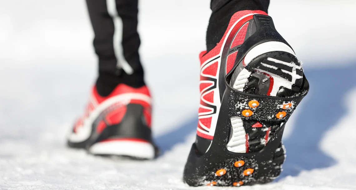Närbild på en persons fötter på som går i snö, iklädda skor med broddar.