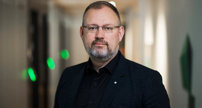 Porträtt av en person i mörk kavaj och svart skjorta som står inomhus. Personen bär glasögon och har kort hår och skägg. Bakgrunden är suddig med ljusa väggar och några gröna ljusdetaljer.