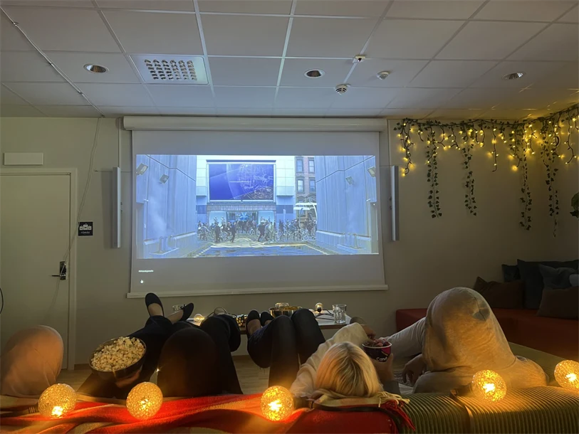 I ett mysigt rum med ljusslingor sitter ungdomar i en soffa och kollar på film på en projektorduk