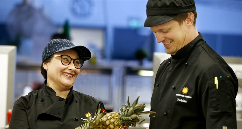 Två kockar i svarta kläder som bär en låda med frukt