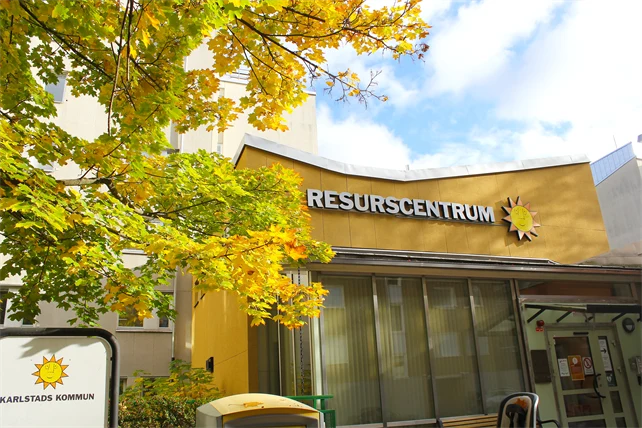Resurscentrums huvudentré med texten resurscentrum och karlstadssolen. Bredvid ingången står ett stort lönnträd med gröngula löv som sträcker sig framåt. Bakom byggnaden syns en klarblå himmel och några vita moln.
