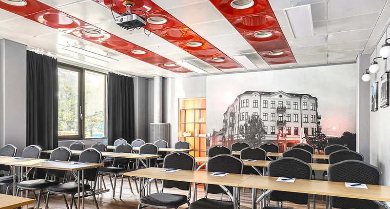 Flera mindre mötesbord med stolar står placerade i ett stort rum med ett vit- och rödrandigt tak.orskärm