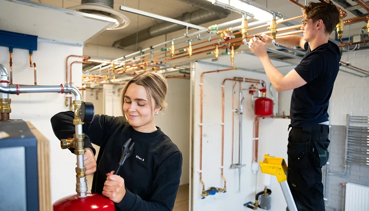 En kille står och drar åt en mutter på ett rör kopplat i taket, med en skiftnyckel. En tjej står bredvid och mekar med en värmepanna.