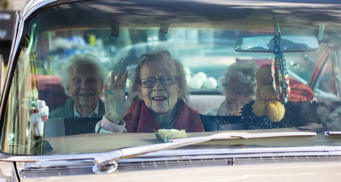 Tre kvinnor i en Cadillac, i förgrunden syns passageraren vinka mot kameran